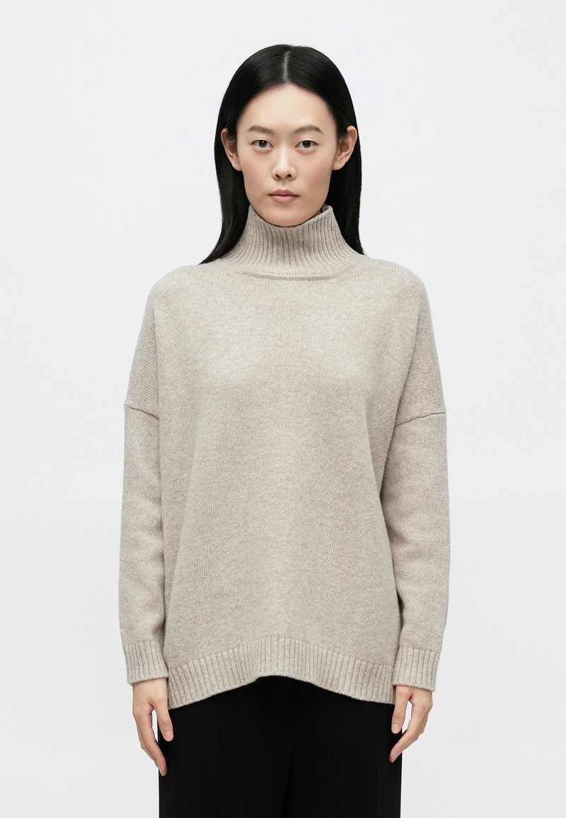 Γυναίκα με ίσια μαύρα μαλλιά, φοράει μπεζ πουλόβερ με ζιβάγκο και μαύρο παντελόνι, στέκεται μπροστά σε λευκό, απλό φόντο.