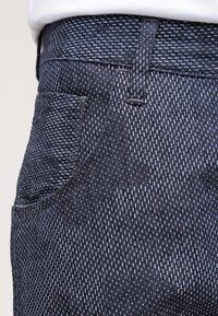 Jeans de mezclilla azul oscuro con un patrón texturizado, que presentan un bolsillo delantero y trabillas para el cinturón, destacando costuras visibles y un corte clásico.