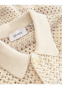 Beige gestricktes Poloshirt mit einer strukturierten, offenen Masche, breitem Kragen und einem weißen Knopf; Etikett trägt "LES DEUX", Größe M.