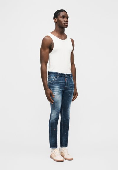 Giovane uomo in piedi, indossa una maglietta bianca senza maniche, jeans aderenti blu strappati, calzini bianchi e sneaker beige, su uno sfondo chiaro uniforme.