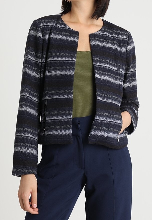 Femme portant une veste zippée à rayures noires et grises sur un haut côtelé vert olive et un pantalon bleu marine, debout avec une main dans la poche.