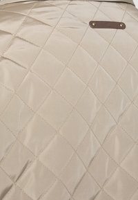 Beige kviltat tyg med ett diamantrutigt mönster, med en brun läderetikett fäst med två metallknappar. Slät, glänsande yta.