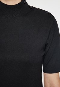 Top scurt tricotat negru cu guler înalt, având o textură ribbed și trei nasturi negri pe umărul stâng.