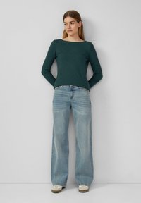 Long-sleeve, dark green top with a boat neck, paired with light blue wide-leg jeans and beige sneakers. Smooth, casual fabric.