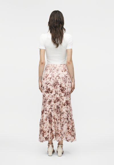 Kvinde med langt brunt hår iført en hvid kortærmet top og en lyserød midikjole med blomster, stående med ryggen til på en ensfarvet baggrund.