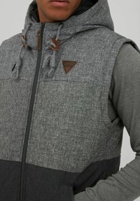 Grauer kapuzen Vest mit strukturierter Oberfläche, mit Reißverschluss vorne, zweifarbiger Gestaltung und einem braunen Lederlogo-Patch. Akzente sind Zugbänder.