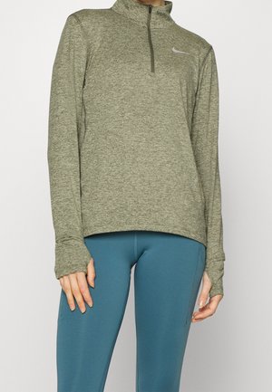 Person trägt einen olivgrünen Langarmpullover mit halbem Reißverschluss und Daumenlöchern sowie eng anliegende, türkisfarbene Leggings.
