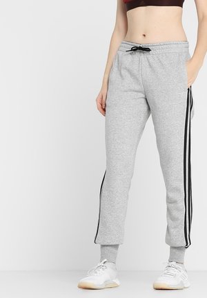 Frau trägt graue Jogginghosen mit schwarzen Seitenstreifen, kombiniert mit weißen Sportschuhen und einem dunklen Sport-BH, der am Bauch sichtbar ist.