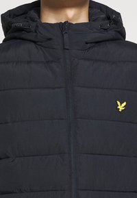 Gilet imbottito blu navy con zip frontale, cappuccio staccabile e cuciture orizzontali; presenta una piccola patch con logo giallo sul petto.