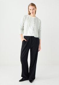 Hellgrau gestrickte Strickjacke mit gewellten Kanten, getragen über einem weißen Hemd und schwarzen weit geschnittenen Hosen, kombiniert mit spitzen schwarzen Schuhen.