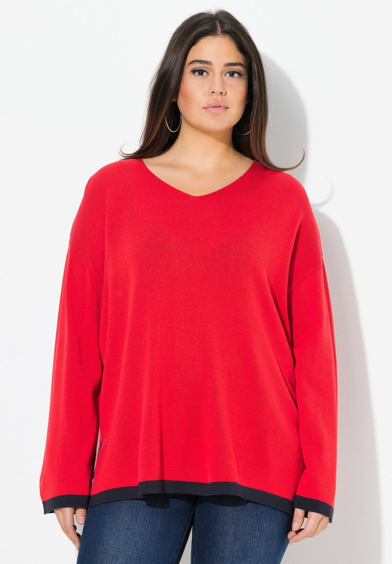 Maglione rosso a V con maniche lunghe e larghe e orlo nero. Realizzato in un morbido tessuto testurizzato. Abbinato a jeans blu scuri.