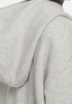 Nærbillede af en person iført en lysegrå hættetrøje, der viser stoftens struktur og sømdetaljer på skulderen og hætten.