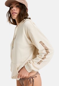 Sudadera beige con un bolsillo tipo canguro, adornada con texto marrón a lo largo de la manga. Combinada con un bikini de parte de abajo marrón. Tela texturizada.