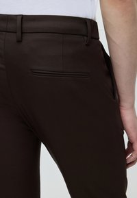 Personne portant un pantalon habillé marron foncé et une chemise blanche, montrant la poche arrière et la main droite reposant sur le côté.