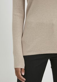 Beige kötött pulóver hosszú ujjakkal és testhez simuló szabással. Puha tapintás és bordázott mandzsetta. Fekete nadrággal párosítva, ami az alján látható.