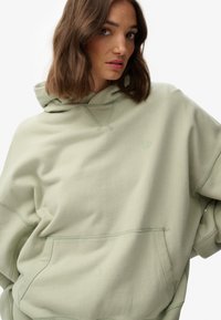 Felpa oversize verde chiaro realizzata in tessuto morbido, caratterizzata da una ampia tasca frontale, dettagli di cucitura sottili e un cappuccio con coulisse.