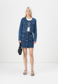 Ženska v modri jeans jakni in krilu z gumbi, ki drži majhno črno torbico in nosi sandale z odprto konico.