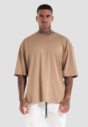 Tetovált férfi laza bézs pólóban és fehér nadrágban, egyszínű világosszürke háttér előtt áll.