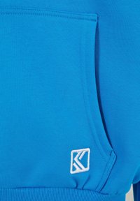Zila džemperis ar priekšējo kabatu, ribotām piedurknēm un baltu izšūtu logo apakšpusē. Mīksts kokvilnas materiāls, gluda tekstūra.