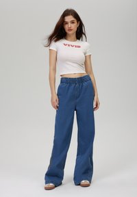 Bijeli kratki crop top s natpisom "VIVID"; plavi široki traperice s elastičnom strukom; model se nalazi uz neutralnu pozadinu.