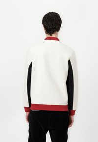 Hvit sweatshirt med rød ribbestrikket krage og hem, svarte sidestriper, teksturert stoff og minimalistisk design. Vist fra baksiden.