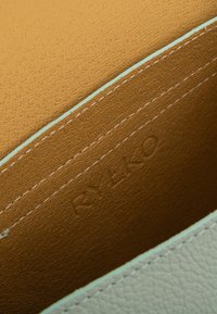 Nahka pussukka, jossa on teksturoitu mintunvihreä ulkopinta ja kontrastinen sileä vaaleanruskea sisus, yksityiskohtana valkoinen ommel ja kohokuvioitu logo.