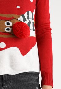 Pull rouge avec le bas blanc, présentant un design de pain d'épice et un détail de pompon rouge. Fabriqué en matériau tricoté. Manches ajustées.