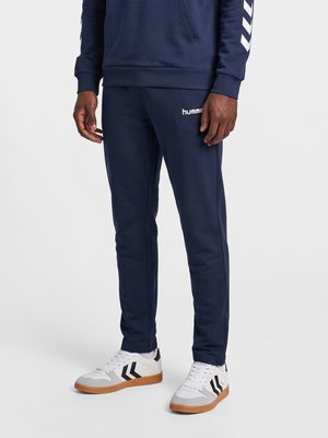 Persona que lleva pantalones deportivos azul marino con el logo "hummel" y zapatillas blancas con rayas de cheurón negras sobre un fondo claro y liso.