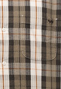 Κοντινό πλάνο πουκάμισου με καρό σε καφέ, μπεζ, μαύρο και άσπρο, με μπροστινή τσέπη και ορατά λευκά κουμπιά.