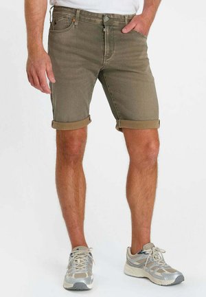 Mann trägt hochgekrempelte olivgrüne Shorts und weiß-graue Laufschuhe, steht mit einer Hand in der Tasche vor weißem Hintergrund.