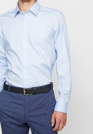 Mężczyzna noszący jasnoniebieską koszulę z długim rękawem, wpiętą w niebieskie spodnie z czarnym paskiem.