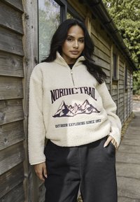 Krämfärgad fleece-pullover med en kvartdragkedja, med en bergsgrafik och texten "NORDIC-TRAIL" i burgundy. Matchad med svarta byxor.