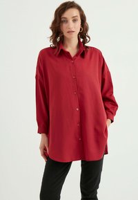 Rotes Langarm-Hemd mit Knopfleiste und Kragen, aus glattem Stoff. Verfügt über einen seitlichen Schlitzsaum und eine lockere Passform. Getragen über schwarzen Hosen.