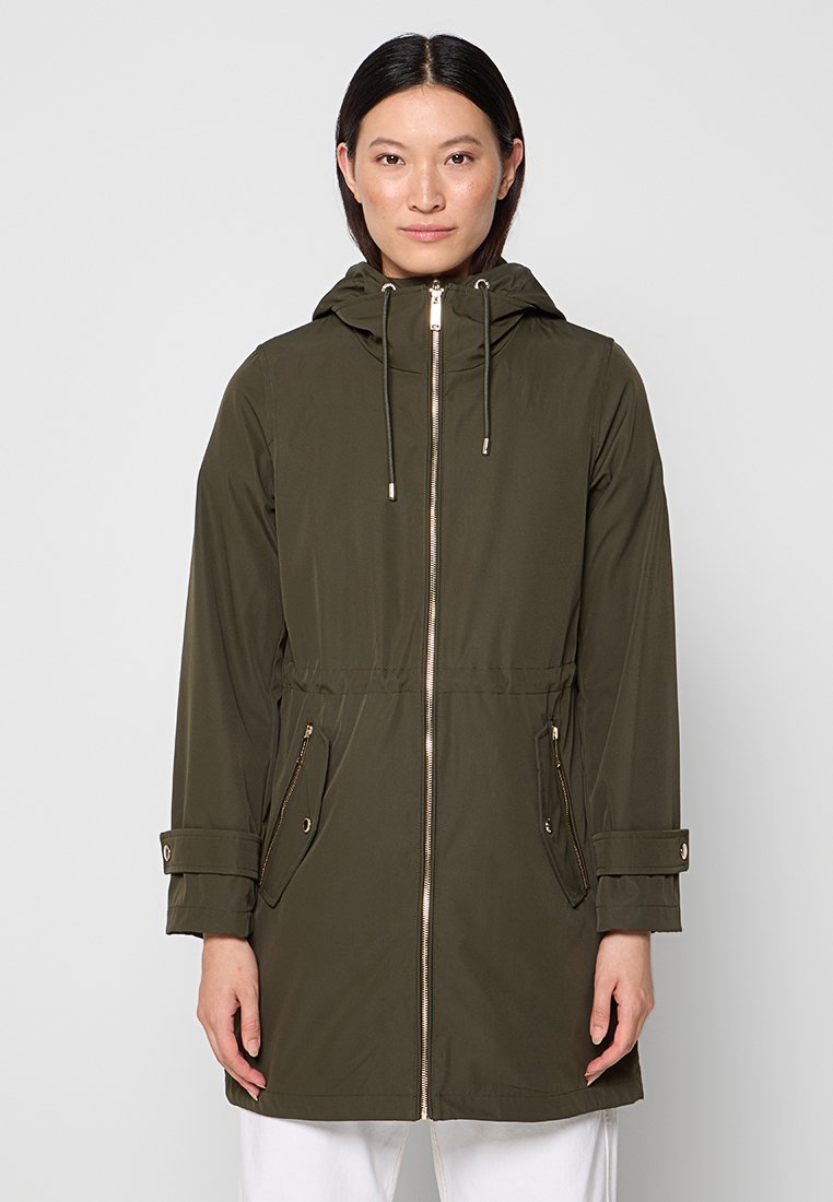 Kobieta w długiej oliwkowo-zielonej kurtce z kapturem, z zamkiem z przodu i zamykanymi kieszeniami, stojąca na tle jasnego, jednolitego tła.