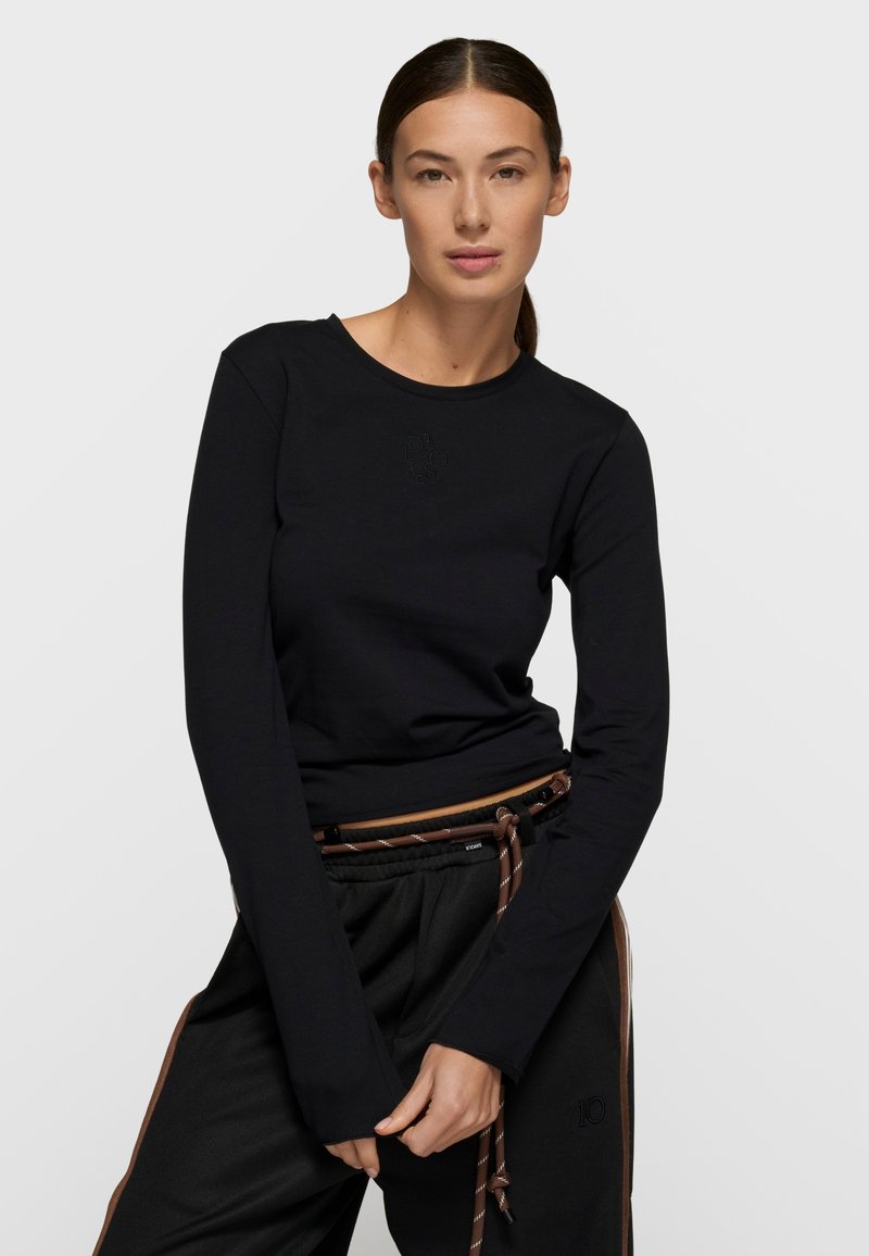 Camiseta negra de manga larga con cuello redondo, hecha de tela suave. Presenta un logo sutil en relieve y mangas entalladas. Se lleva con pantalones negros.