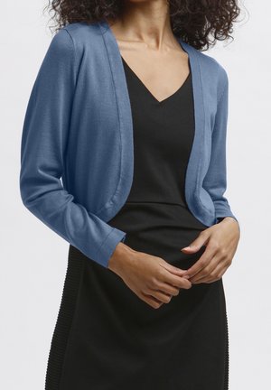 Frau in einem enganliegenden schwarzen Kleid mit einem offenen, kurz geschnittenen blauen Cardigan, die Hände locker vor der Taille verschränkt.