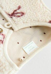 Beige Fleece-Babybekleidung mit einer weichen, strukturierten Außenseite. Die Innenseite zeigt einen cremefarbenen Stoff mit burgunderfarbenen Punkten und einem Markenetikett.