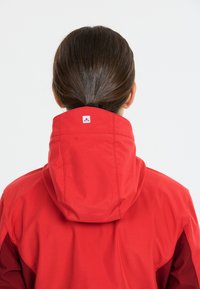 Red waterproof jacket with a high collar and hood. Features a logo on the back, reflective detailing, and a smooth texture.