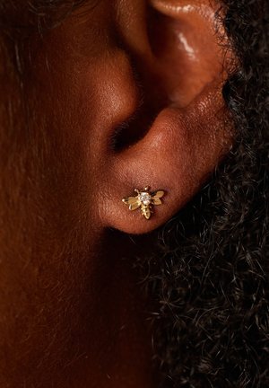 Gouden bij-vormige oorring met een kleine heldere edelsteen in het midden, liggend op een donkere huidtint, met getextureerd haar op de achtergrond.