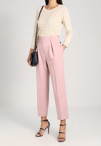 Beżowa bluzka z długim rękawem o teksturowanym wzorze, zestawiona z różowymi spodniami o wysokim stanem i przezroczystymi szpilkami. W zestawie torebka na ramię w kolorze czarnym.