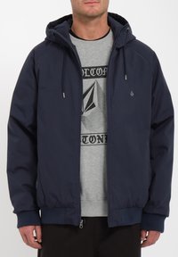Donkerblauwe ritsjack met capuchon, voorzien van ribbels aan de mouwen en de onderkant, gedragen over een grijze sweatshirt met opdruk. Soepele textuur, minimale details.