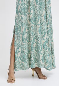 Dlhé, vzorované šaty s svetlou krémovou základnou a teal paisley vzorom. Obsahujú bočný rozparok, kombinované s béžovými sandálmi na vysokom opätku.