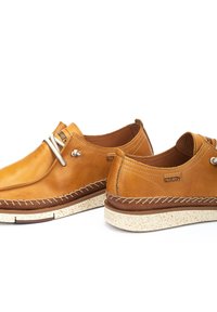 Zapatos de cuero marrón claro con costuras blancas, punta redondeada y suela de goma con un diseño moteado. Diseño minimalista con acentos metálicos.