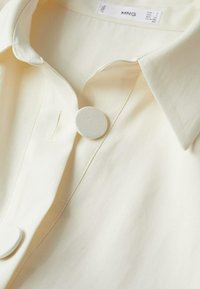 Chemise couleur crème avec un col classique, une texture lisse, et un grand bouton rond à l'avant, avec une étiquette d'entretien du tissu visible.