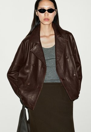 Woman wearing dark brown leather jacket, gray tank top, black skirt, black sunglasses, and holding a black handbag against plain background.