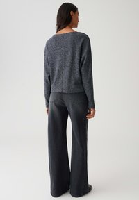 Femme portant un haut long à manches gris foncé texturé et un jean noir délavé à jambes larges, se tenant de dos devant un fond uni.