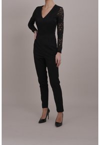 Czarny koronkowy body z głębokim dekoltem w serek i długimi rękawami, w zestawieniu z czarnymi spodniami o wysokiej talii i zwężanym kroju oraz czarnymi szpilkami.