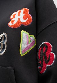 Felpa nera con colorati toppe ricamate di forme diverse: "H" arancione, cuore verde e rosa, "B" grigia e "R" rossa.
