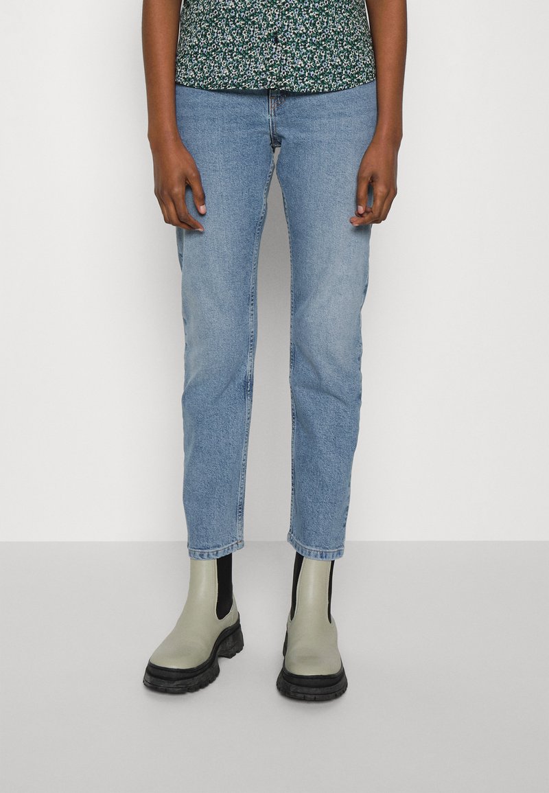 Πλενόμενα μπλε τζιν με ίσια γραμμή, ελαφριά φθορά στα πόδια, συνδυασμένα με πράσινες μπότες αστραγάλου με μαύρες καουτσούκ σόλες.