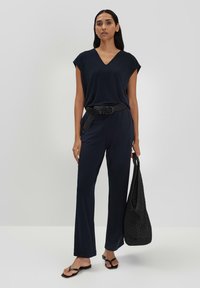 Navyblauwe jumpsuit met een V-hals, korte mouwen en een geweven riem. Gecombineerd met een zwarte totebag en zwarte sandalen. Glad, lichtgewicht stof.